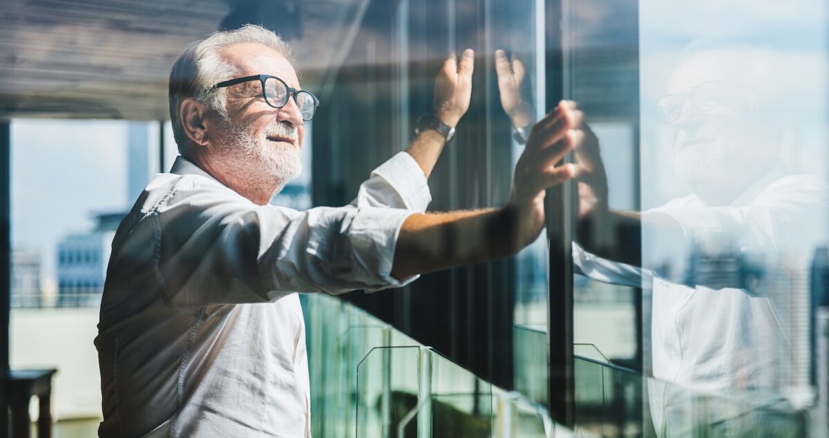 Senior male business owner standing by a window in a modern office, reflecting on retirement planning, symbolizing the risks of relying solely on a business to fund retirement.