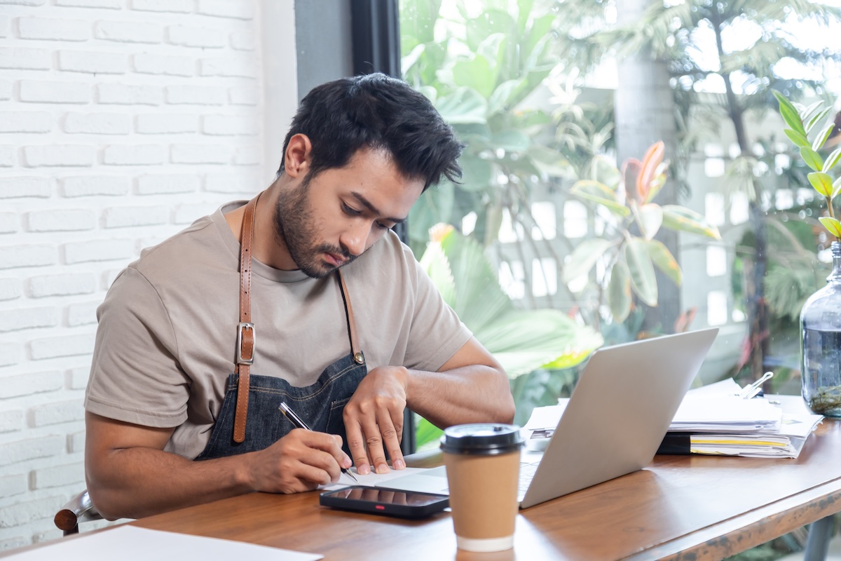 Small business owner in casual attire and apron calculating finances at a desk with a laptop, documents, calculator, and coffee; representing proactive tax planning strategies for entrepreneurs.