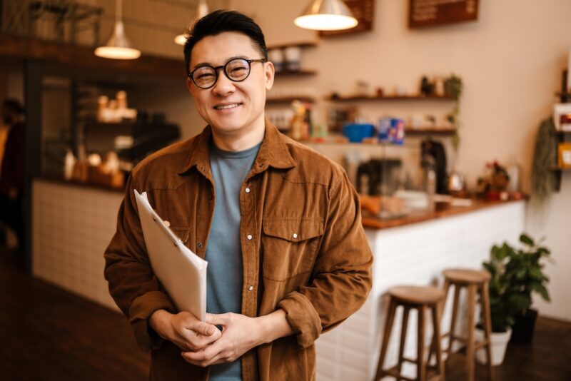 Smiling Buffalo Grove small business owner standing in his café holding financial documents, representing smart financial planning and retirement strategies for entrepreneurs.